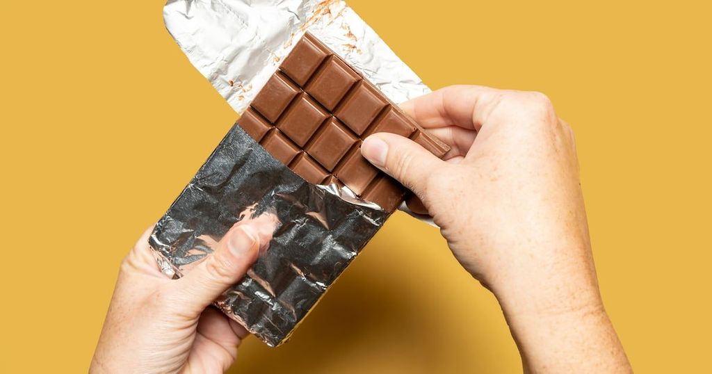 A hand is holding a piece of chocolate bar wrapped in foil against a golden background.