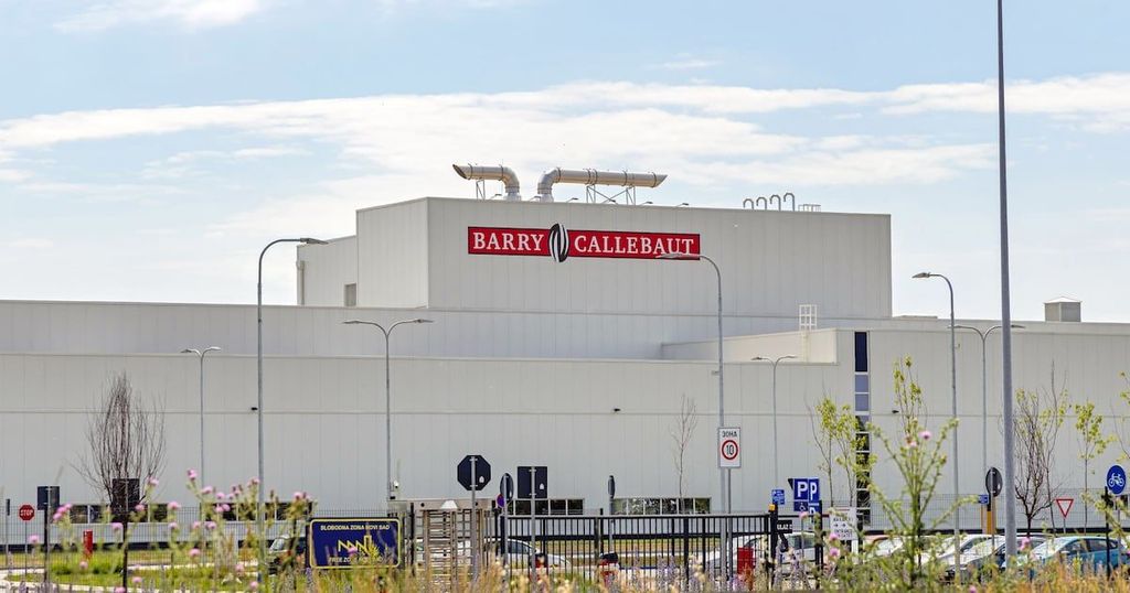 A large, industrial building with the Barry Callebaute logo is viewed against a cloudy sky, featuring parking areas and streetlights.