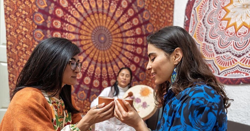 Two women exchange cups of tea in front of intricately patterned Indian tapestries.