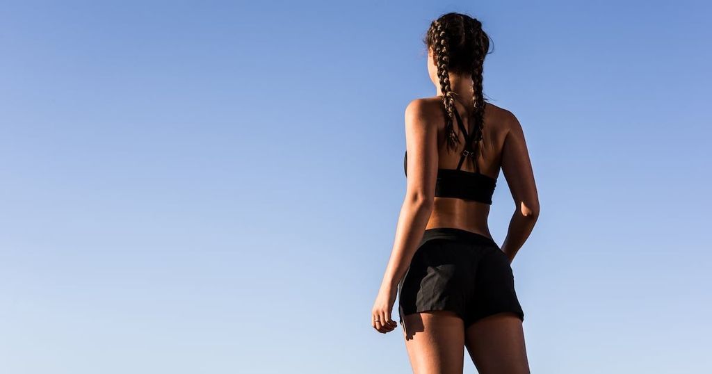 A woman with braided hair stands with her back to the viewer, wearing a black sports bra and shorts against a bright blue sky.