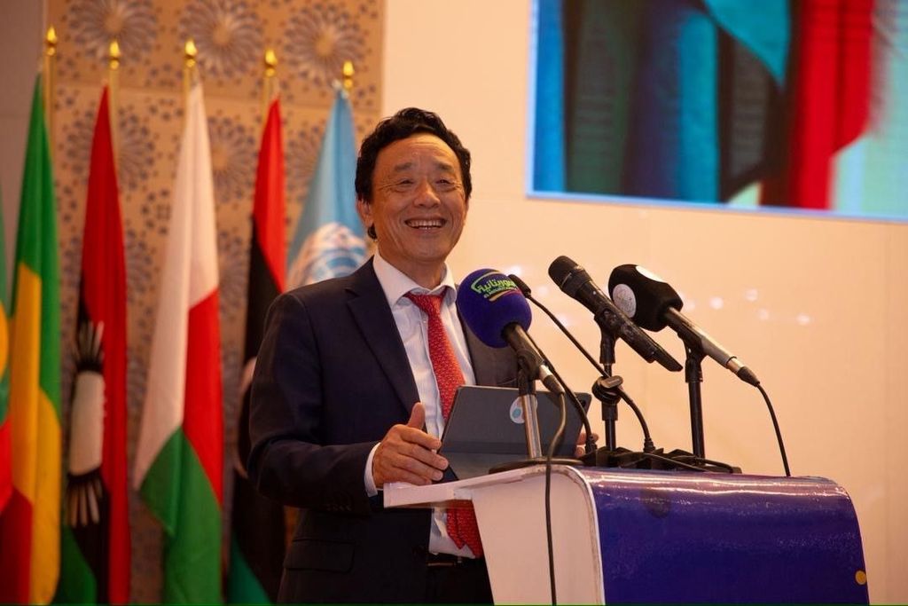 A man in a suit is speaking at a podium surrounded by flags and microphones during a formal event.