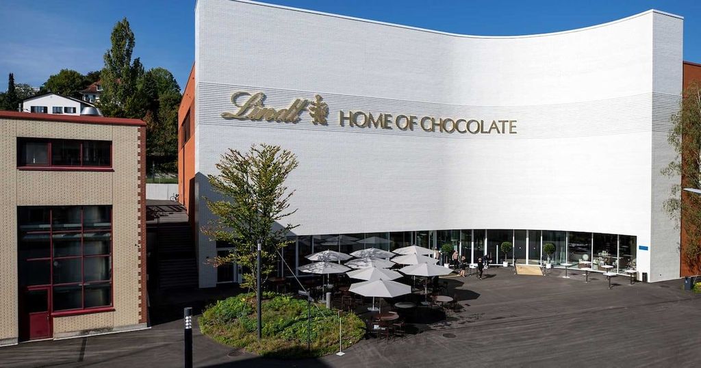 A large, white building with the “Lindt HOME OF CHOCOLATE” sign is flanked by smaller buildings and features an outdoor seating area with tables and umbrellas.